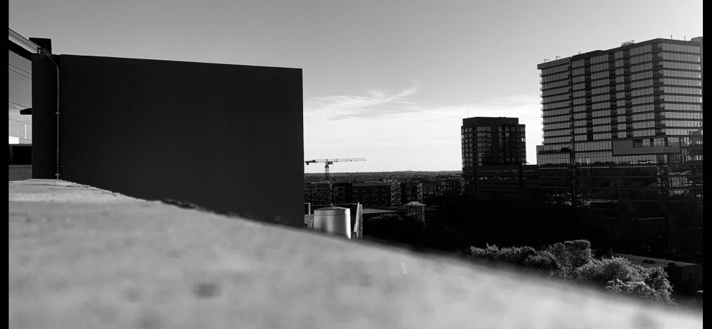 A crane operating between several high-rise building in a black and white filter with the tops of some trees at the bottom right.